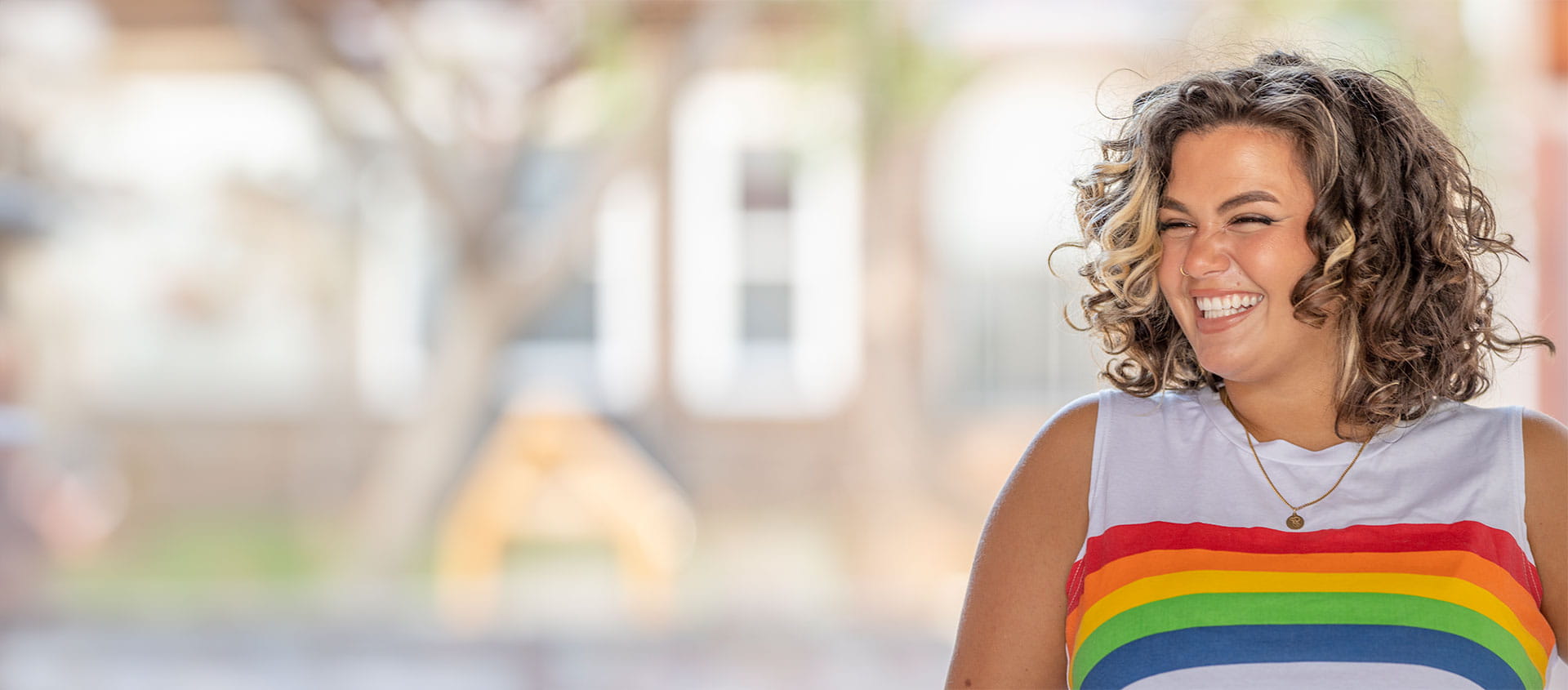 Une personne vêtue d'un t-shirt sans manches orné d'un motif arc-en-ciel, symbole d'inclusion et de fierté, se tient debout à l'extérieur et sourit.