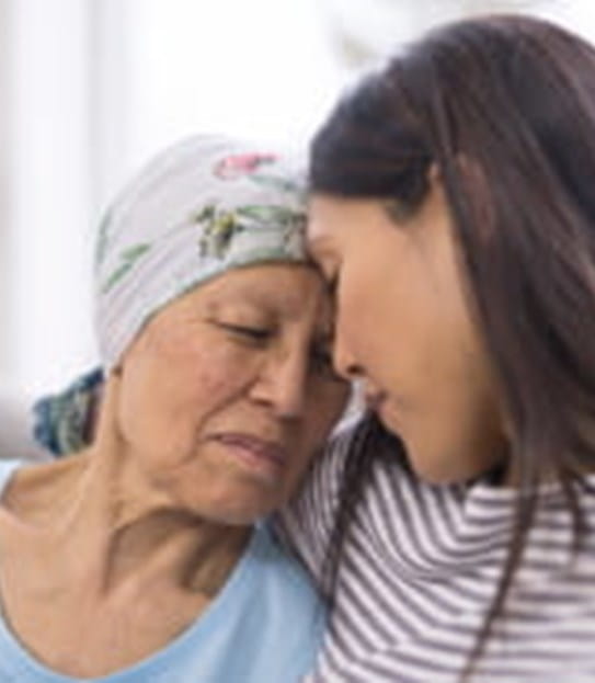An elderly woman with cancer and wearing a head covering is embracing her adult daughter. They are sitting on a couch and their foreheads are touching.