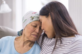 An elderly woman with cancer and wearing a head covering is embracing her adult daughter. They are sitting on a couch and their foreheads are touching.