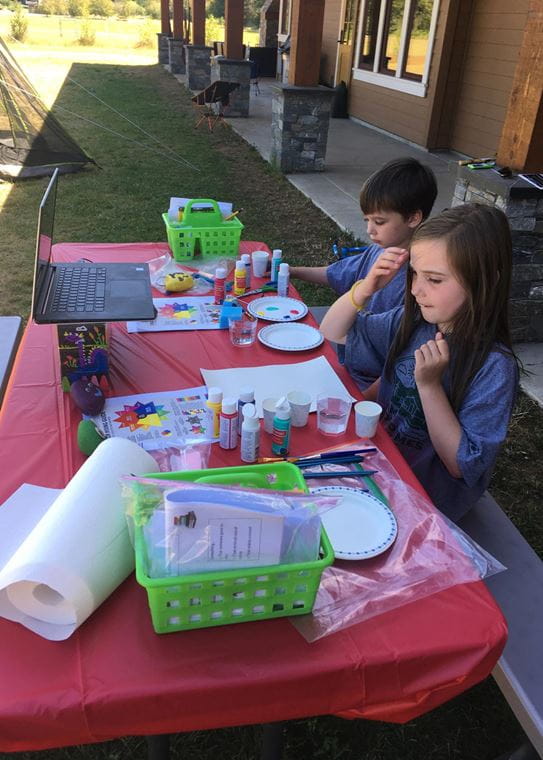 Two children sit at a park bench covered with an assortment of craft supplies