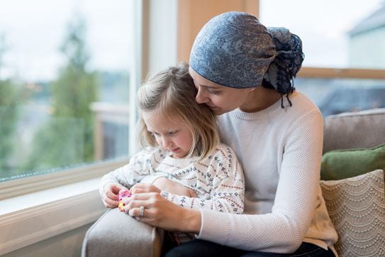 A beautiful young woman with cancer holds her preschool-age daughter in her lap by their living window. They are holding and playing with a little plastic toy and mom is smiling. She is also wearing a headscarf.