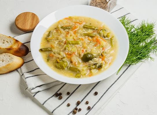 Un bol de soupe à l’orge et aux légumes est déposé sur une serviette, près de quelques tranches de pain et de fines herbes.