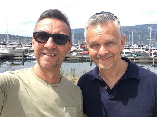 Two men stand smiling beside each other with boats in the background