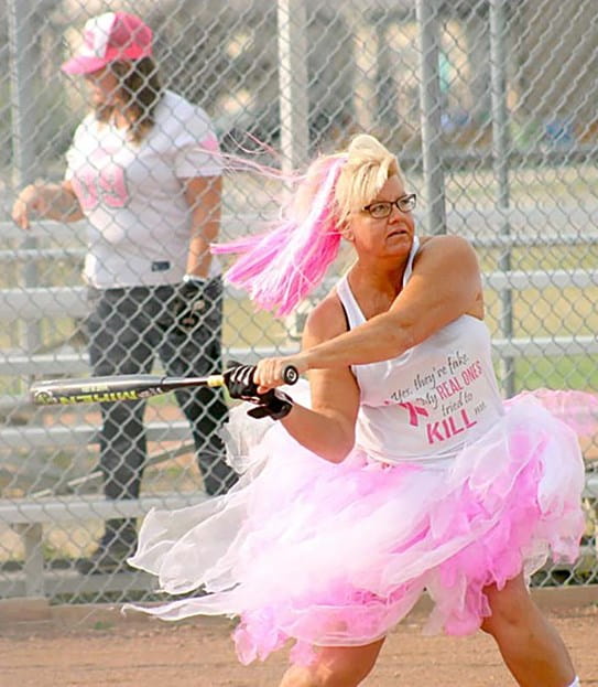 A woman in a large pink skirt with pink dyed hair swinging a baseball bat