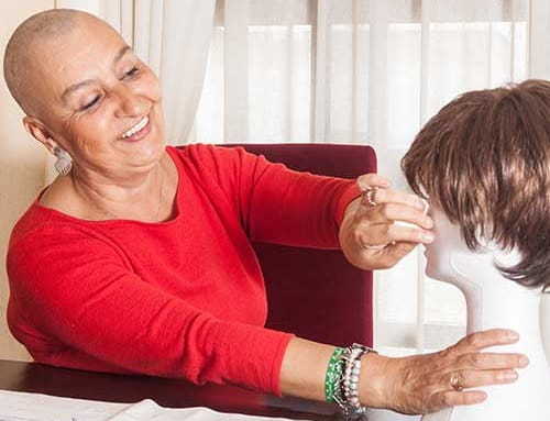 A bald woman smiles as she adjusts a wig on a wig stand