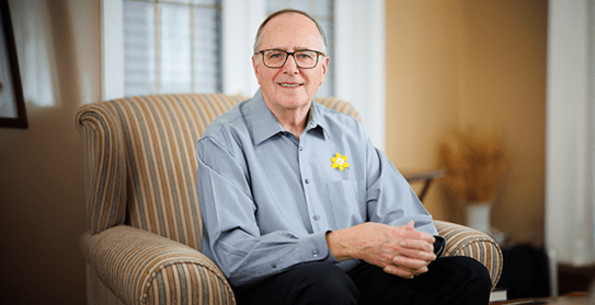 Photo of Allen Goudie wearing a daffodil pin on his shirt, sitting in an armchair.