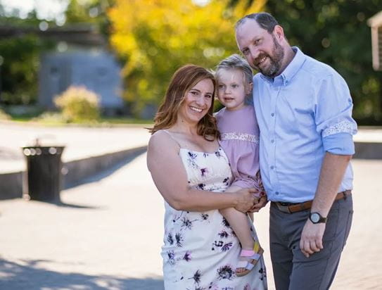 2. Natalie, souriante, debout à côté de son mari et tenant leur enfant