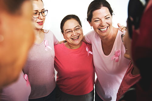 A small group of women wearing pink t-shirts and a pink ribbon have their arms around each other’s shoulders.