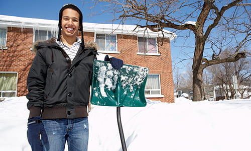 A young man is smiling and holding a snow shovel with snow behind him.