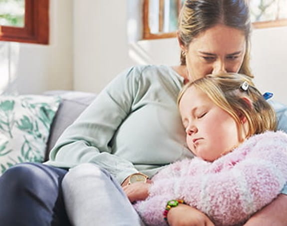 A mother and her young daughter are sitting on a couch. The mother is kissing the top of her daughter’s head while her daughter sleeps. 