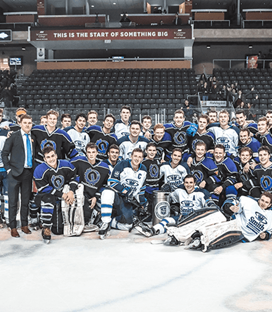 Les équipes de hockey des facultés de commerce et de génie de l’Université Queen’s se tiennent sur une patinoire avec le comité organisateur de Cure Cancer Classic.