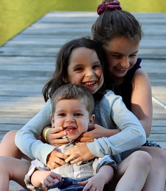 Three siblings sitting in a row hugging each other and smiling.