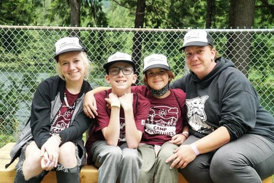 Trois campeurs assis à gauche de leur mère. Tous sourient et portent le chandail et la casquette du Camp Goodtimes.