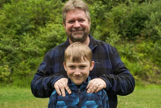 A little boy smiling straight at the camera. His father is standing behind him smiling with his hands on his shoulders.