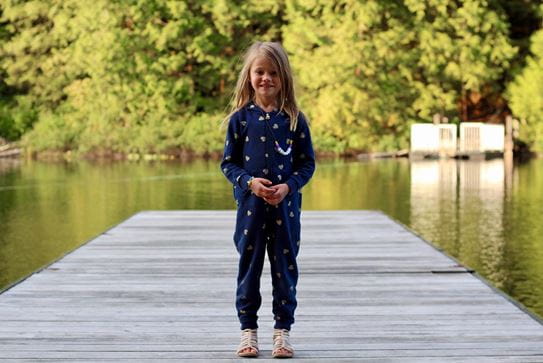 A little girl in a onesie smiling and standing on a dock at a lake.