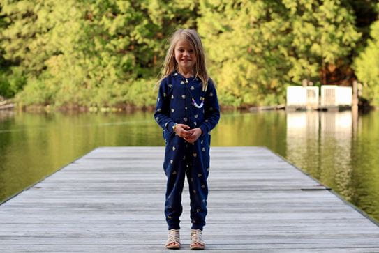 A little girl in a onesie smiling and standing on a dock at a lake.