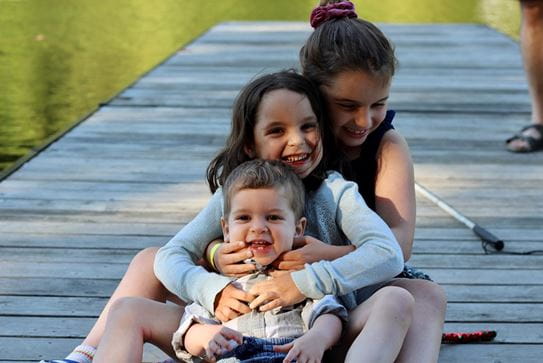 Deux sœurs et un frère assis l’un derrière l’autre en s’étreignant et en souriant.
