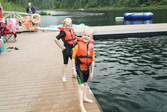 Two smiling siblings wearing lifejackets and standing on the side of a dock.