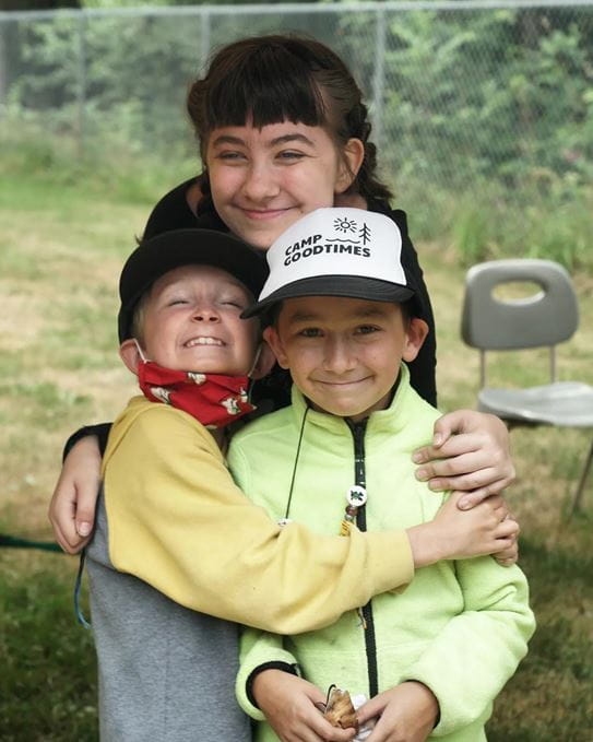 Three kids sitting in a row on a dock smiling with their arms around each other.