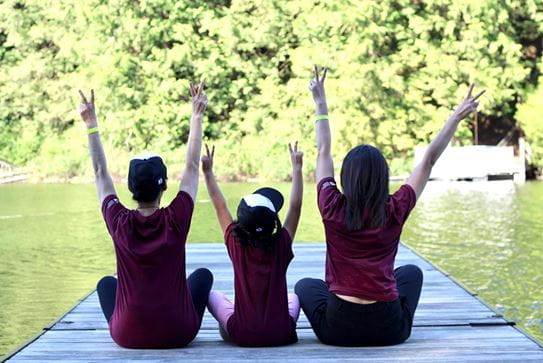Three campers with their backs to the camera sitting on a dock. Their arms are up and they’re making peace signs.