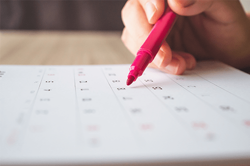 Une main tenant un crayon rose devant un calendrier apposé sur une table