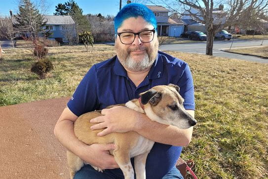 Roy sitting on the step of his home holding his dog. He has blue dyed hair and is wearing blue clothes.