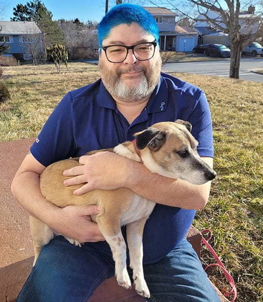 Roy sitting on the step of his home holding his dog. He has blue dyed hair and is wearing blue clothes.