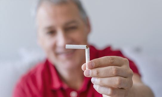 A blurred-out older man holding up a broken-in-half cigarette that is in focus.