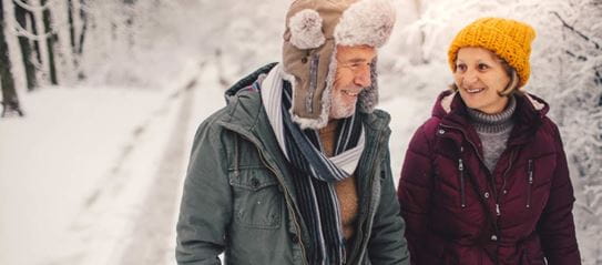 An older couple smiling and walking down a snow-covered forest trail.