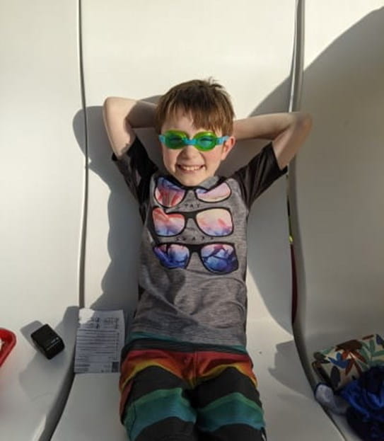 Linden wearing beach clothes and sitting on a bench with his hands behind his hand.
