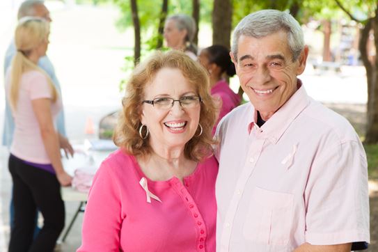 An older couple wearing pink ribbons embracing and smiling.  