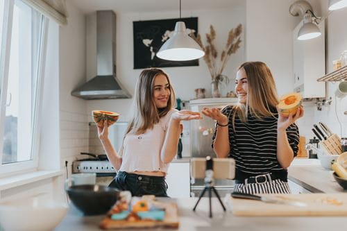 Deux femmes se sourient alors qu’elles se filment en train de faire à manger dans une cuisine.