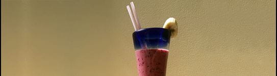 A fruit smoothie in a glass with a slice of banana on the rim. 