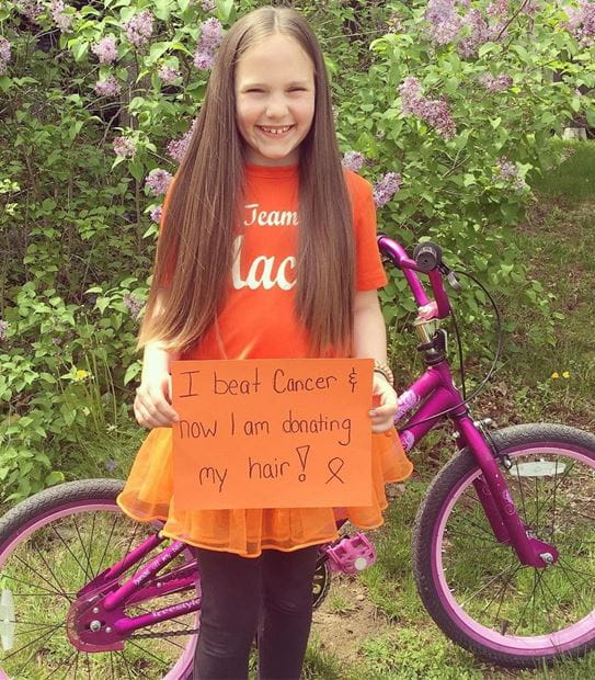 Maci Millen in front of her bicycle holding a sign that says I beat cancer & now I am donating my hair.
