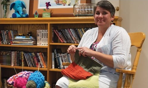 A person in a chair, holding knitting needles in front of a bookcase.