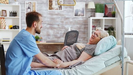 A person in scrubs comforting a person in a hospital bed at home.