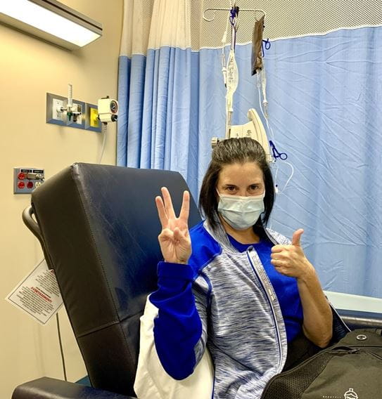 Jennifer, sitting in a chair in a hospital receiving chemotherapy treatment. She’s holding up three fingers with her right hand and a thumbs up with her left hand.