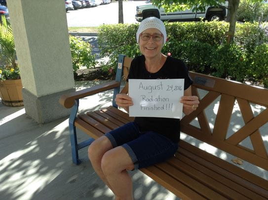 Vickie Iachetta, sitting on a park bench, holding up a sign that reads “August 24, 2016 Radiation Finished!!”