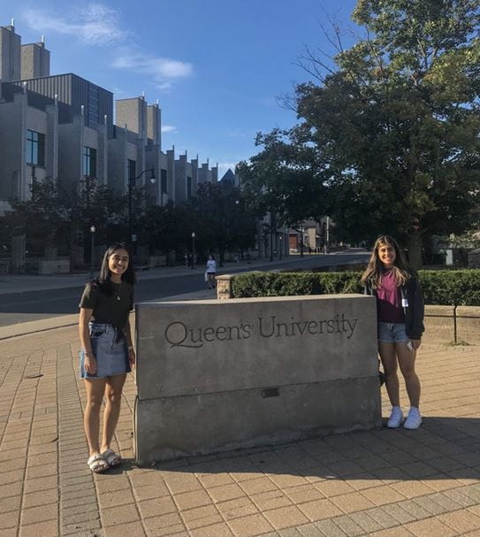 Alaina and Jillian standing outside on opposite sides of a stone that says, “Queen’s University.”