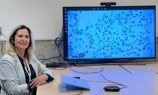 Dr April Khademi sitting at a desk with a laptop and a large monitor showing cells