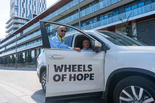 James, standing at the front passenger entrance of a Wheels of Hope car, helping a passenger get out.