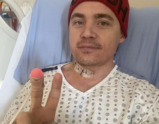 Shawn laying in a hospital bed wearing a hospital gown, holding a bottle and making the peace sign.