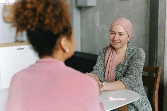 A person wearing a headscarf, sitting at a kitchen table and smiling at another person who has their back to the camera.