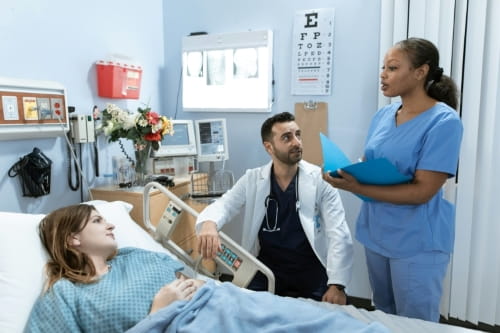 Two doctors speaking to a person in a hospital bed. One doctor is standing and holding a chart, and the other is sitting by the bed.