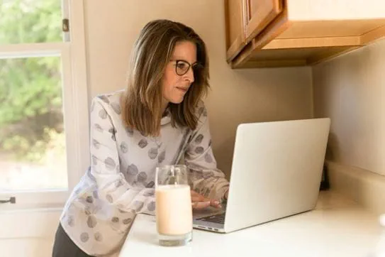 A person standing at a counter with a laptop and a glass of milk in front of her.