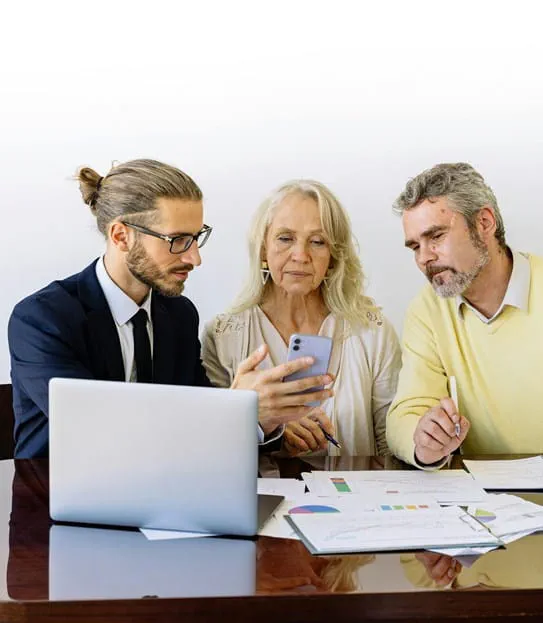 A man and woman are sitting at a table with financial documents in front of them while a man with a laptop shows them something on his cell phone.