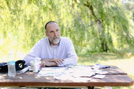 Angus Pratt assis à une table sous les arbres, les bras croisés.