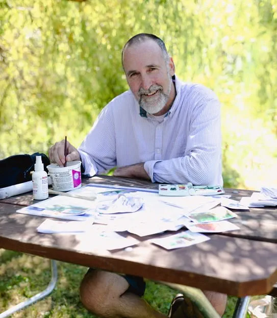 Angus Pratt sitting at a table underneath trees with his arms crossed.