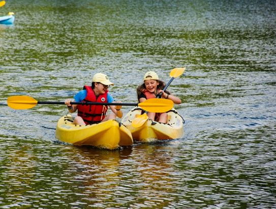 Two children wearing lifejackets while paddling kayaks in the lake and smiling.