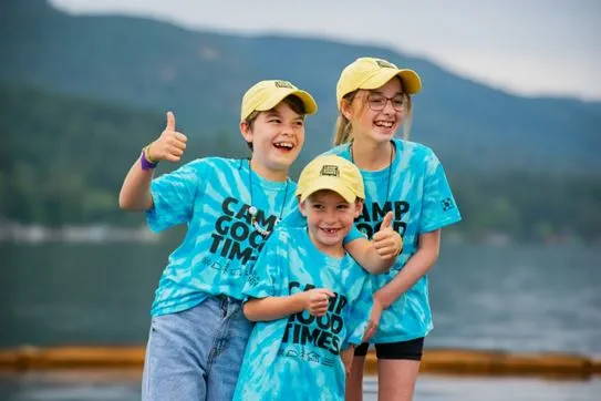 Three kids wearing matching Camp Goodtimes shirts and hats and smiling.